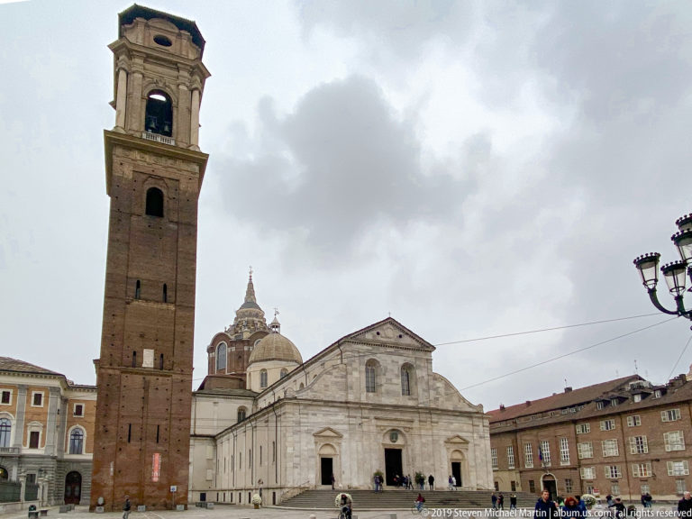 Wide frontal view of Turin Cathedral beside the brick bell tower, with the Shroud Chapel dome rising behind.