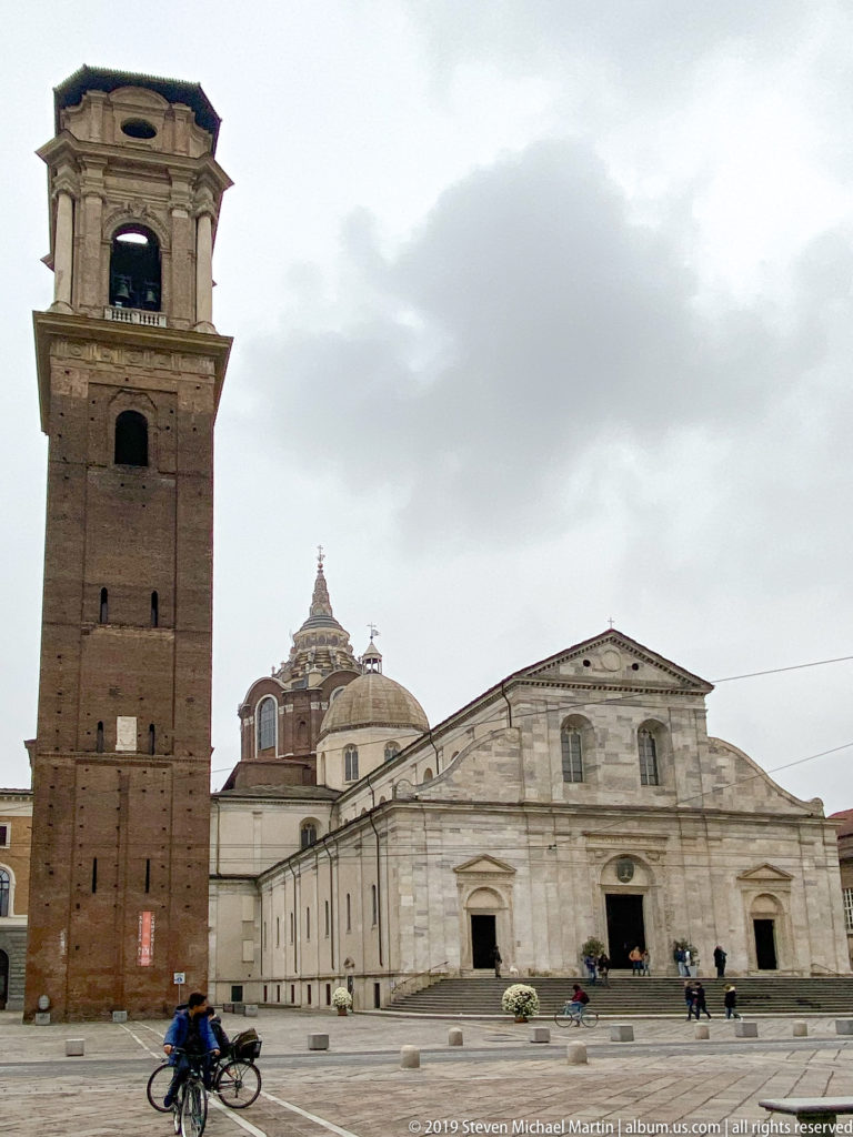 Cathedral of Saint John the Baptist and its tall brick bell tower seen across the piazza beneath an overcast sky.