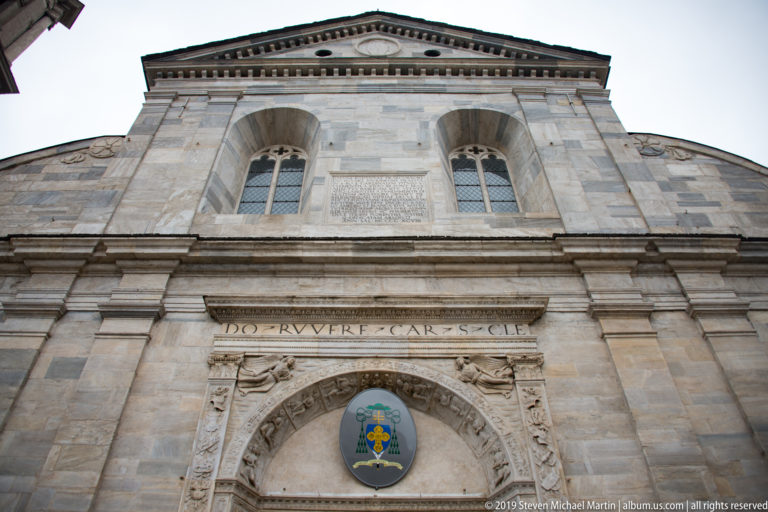 Low-angle detail of Turin Cathedral’s pale stone façade above a portal, including carved inscription, crest, and paired upper windows.