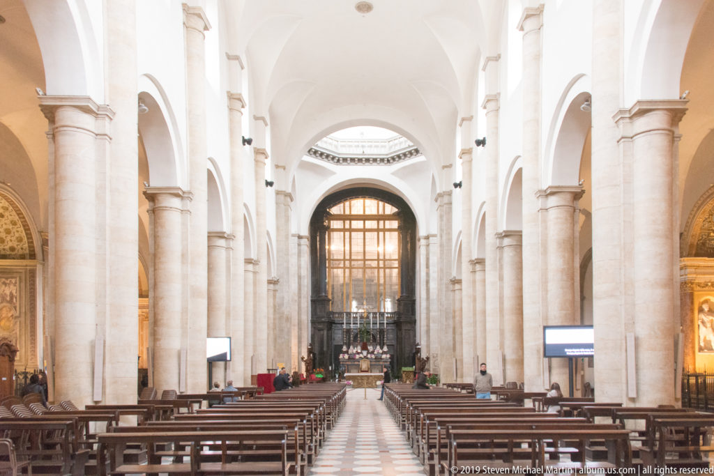 Bright cathedral nave with repeated stone arcades, white vaulting, pews, and the sanctuary centered in the distance.