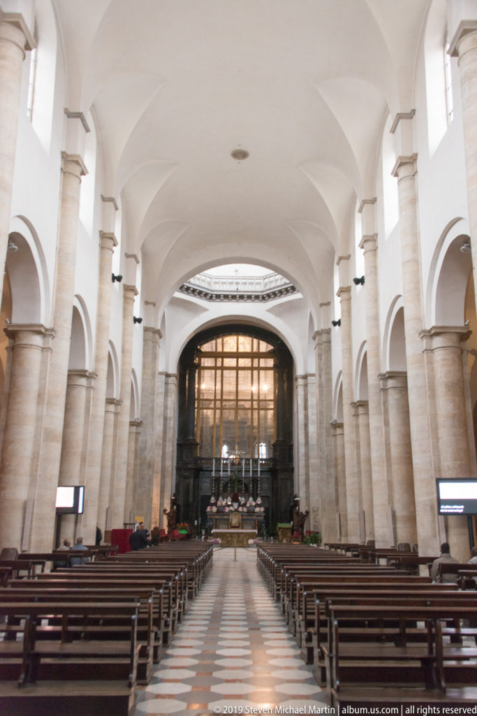 Central aisle of Turin Cathedral lined with pale stone columns and wooden pews, looking toward the screened sanctuary.