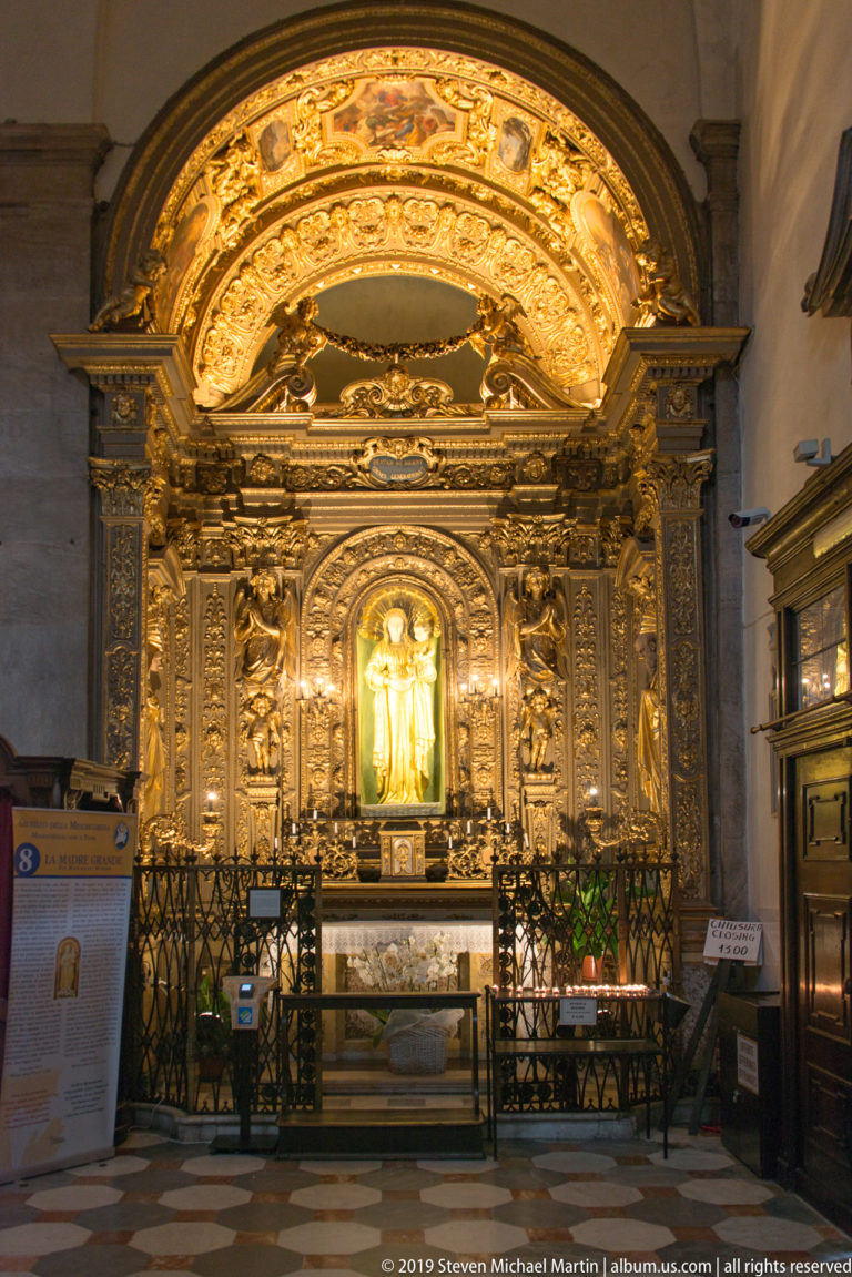 Highly gilded chapel altar centered on a brightly illuminated standing sacred figure behind a decorative metal rail.