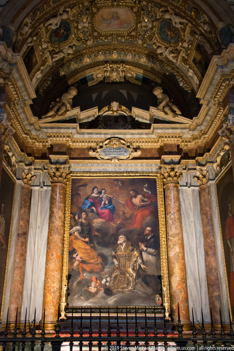 Large multi-figure religious painting set between colored-marble columns beneath an ornate gilded chapel vault.