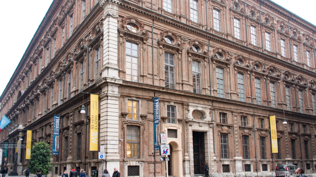 Oblique street view of the Museo Egizio in the former Collegio dei Nobili, with rusticated façade, tall windows, and yellow museum banners.