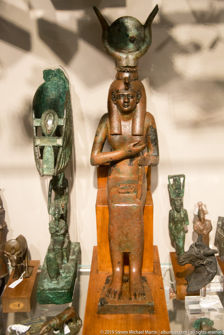 Group of small Egyptian deity statuettes in bronze and other materials, including a central reddish figure with a tall horned headdress.
