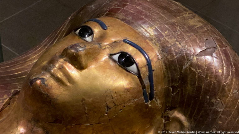 Close view of a gilded Egyptian coffin face with large inlaid eyes, blue cosmetic lines, and a striped wig.