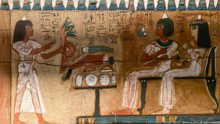 Painted Egyptian funerary panel with hieroglyphs, standing and seated figures, and an offering table filled with vessels and food.