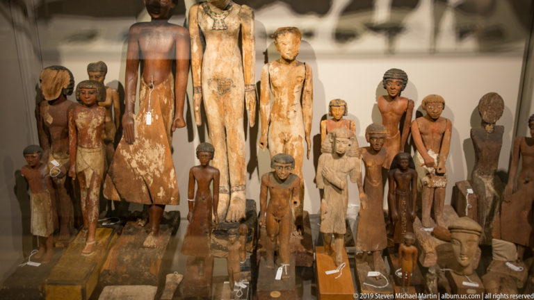 Group of ancient Egyptian painted wooden figures of varied sizes, with dark wigs, white garments, and worn surfaces.