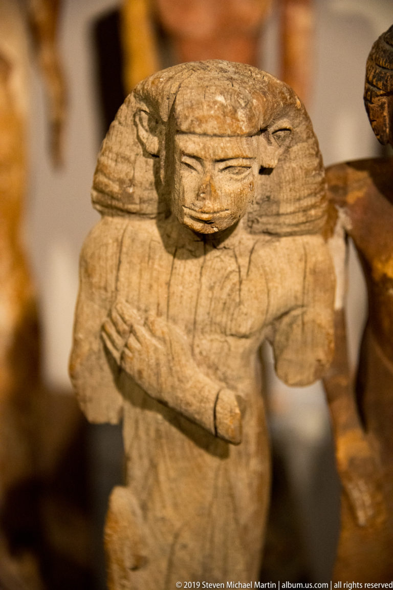 Small worn wooden Egyptian figure with a broad layered wig, rounded face, and both forearms crossed over the chest.