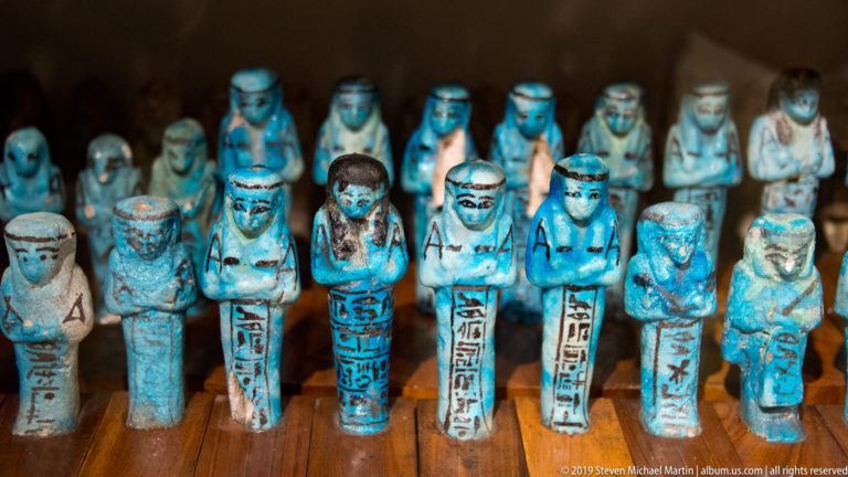 Rows of blue faience shabti figurines with mummiform bodies, crossed arms, wigs, and dark hieroglyphic inscriptions.