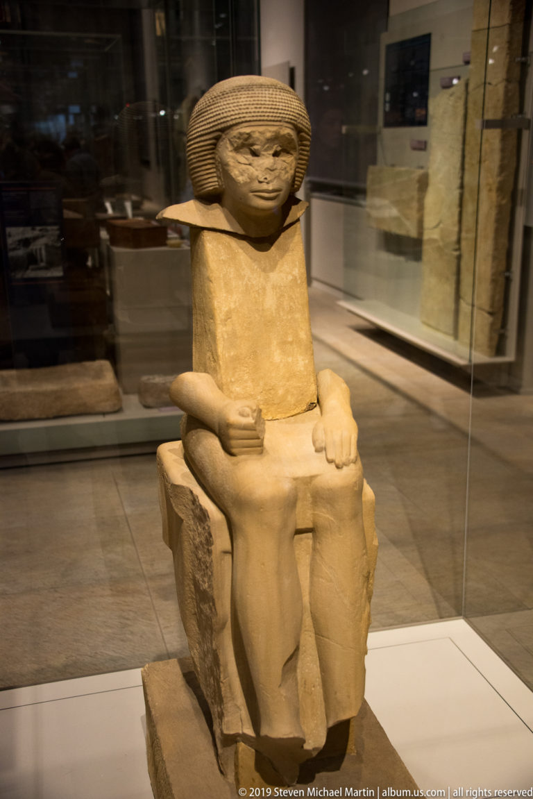 Full seated statue of an Egyptian man with broad striated wig, simple garment, hands on thighs, and feet forward.