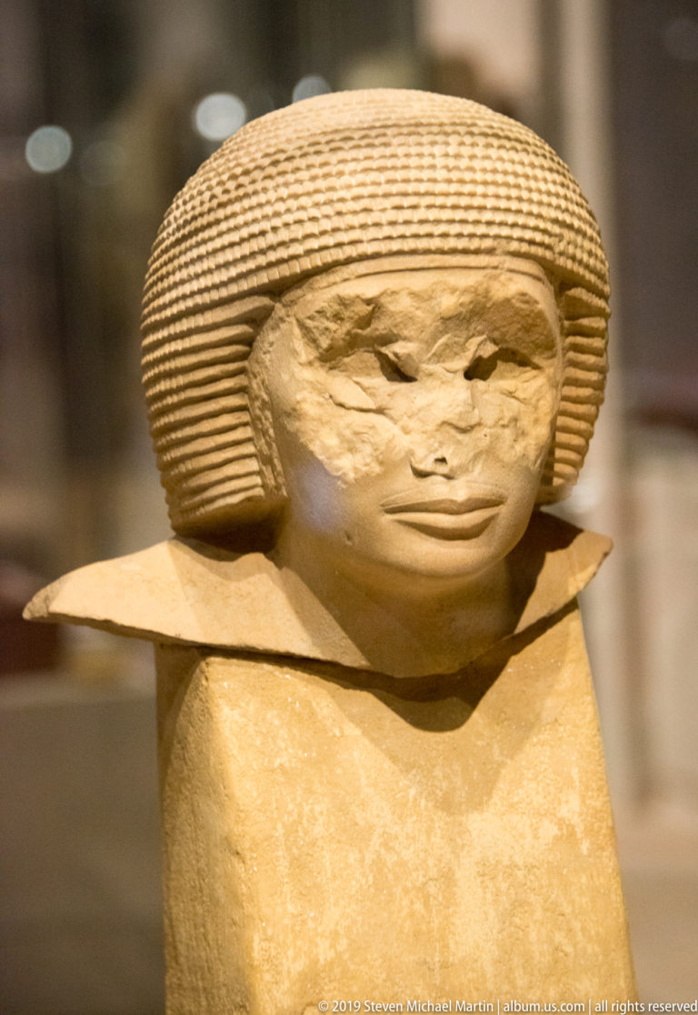 Pale Egyptian male head with broad horizontally striated wig, softly modeled face, and truncated neck on a museum mount.