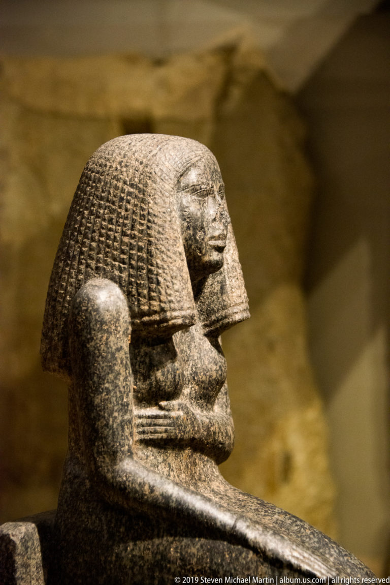 Profile view of a dark stone seated Egyptian figure with textured wig, crossed arms, and compact block-like body.