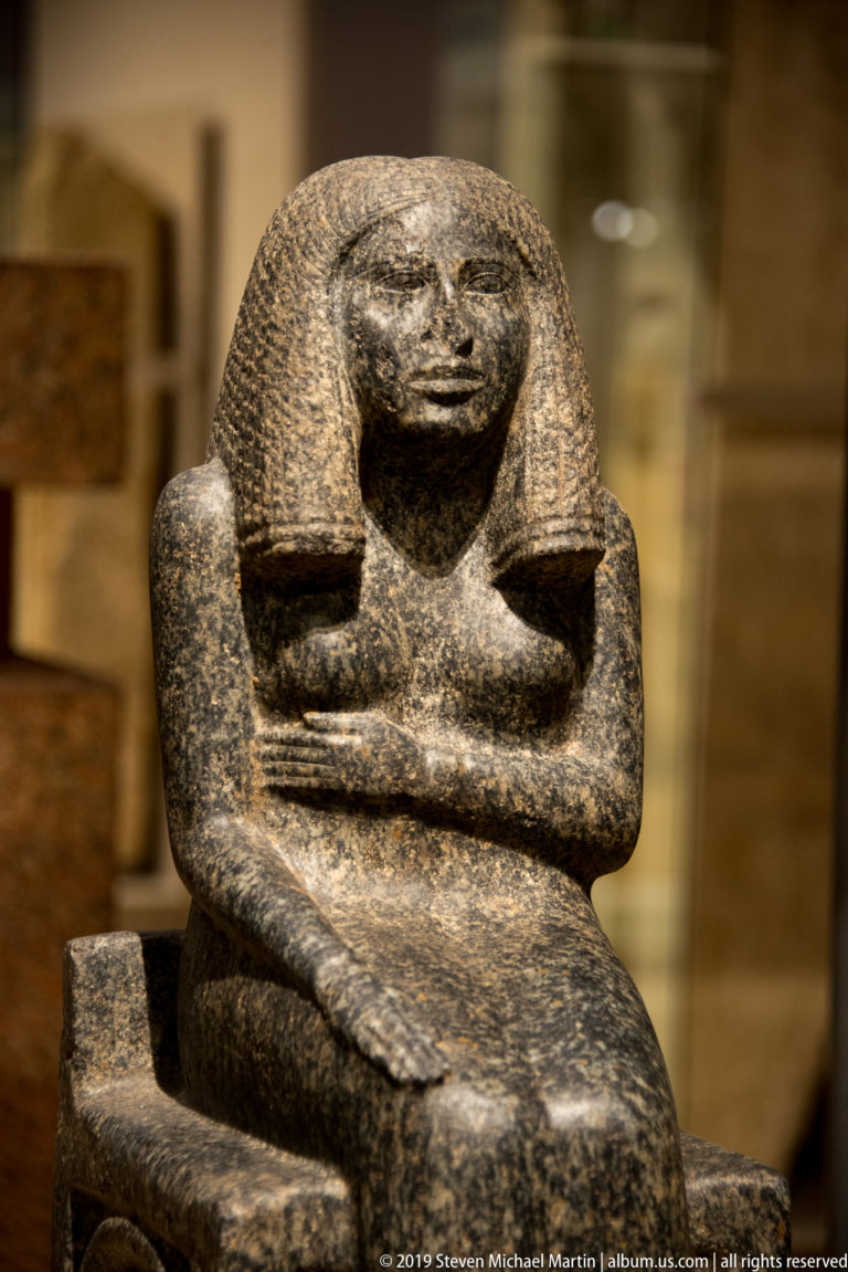 Dark stone seated Egyptian figure with broad textured wig, arms crossed over the knees, and a compact block-like pose.