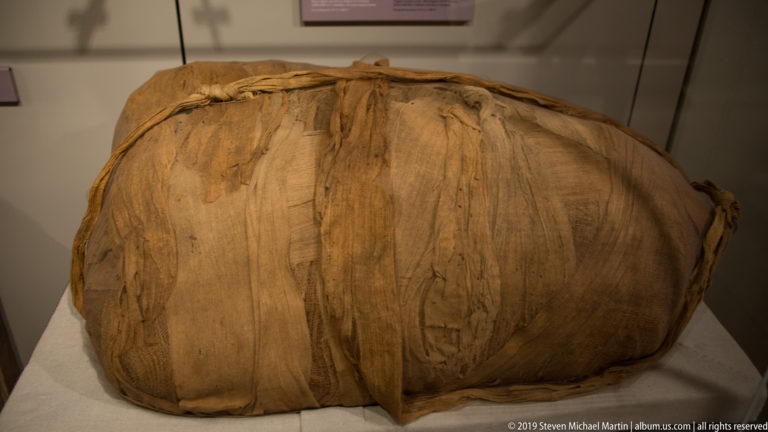Large oval Egyptian funerary bundle wrapped completely in overlapping layers of brown linen with gathered seams.