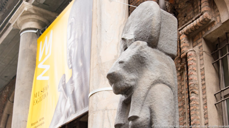 Large weathered Egyptian stone figure beside a yellow Museo Egizio banner, framed by pale stone columns and brick architecture.