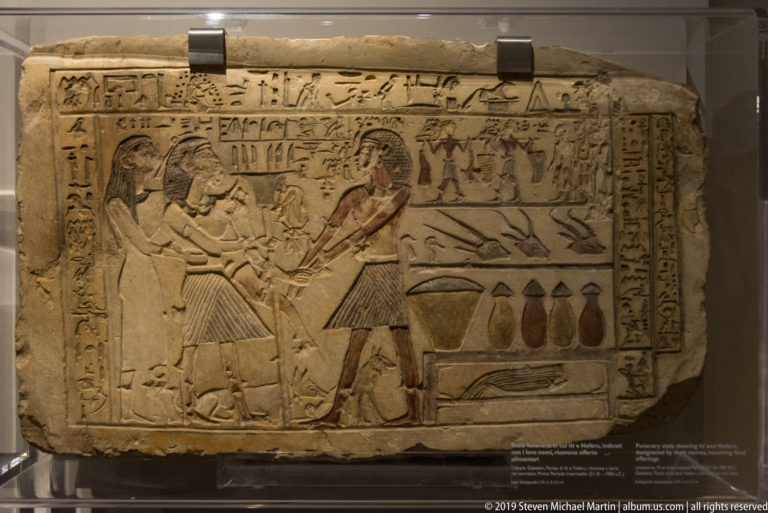 Painted Egyptian funerary stela with a seated couple, standing attendants, hieroglyphs, and rows of food and vessels.