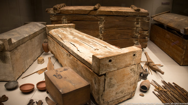 Wide display of plain wooden coffins, boxes, pottery, reed or arrow shafts, and assorted burial goods arranged together.