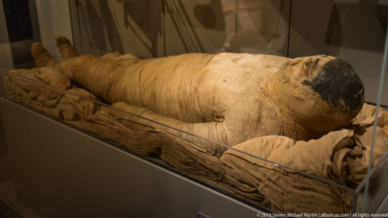 Full-length human mummy tightly wrapped in pale linen with a darkened exposed face, resting in a shallow textile-lined support.