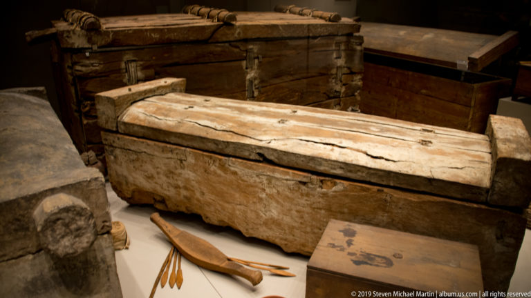 Group of plain ancient wooden coffins with thick plank lids and weathered surfaces, displayed with smaller burial objects.