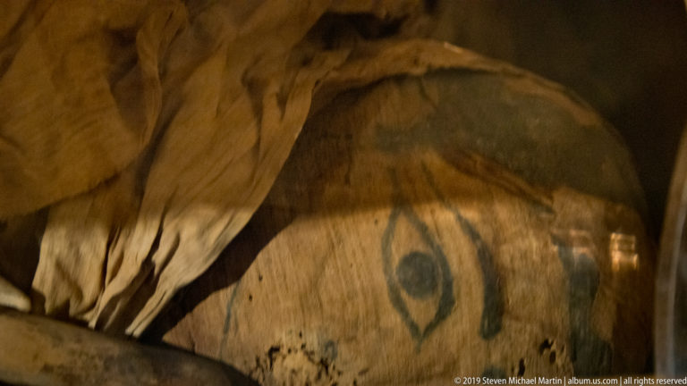 Close detail of a large painted eye on a brown mummy covering or funerary mask with surrounding wrappings and surface wear.