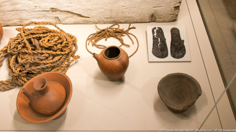 Display case with coiled rope, ceramic vessels, dark bowl, and a pair of ancient sandals or sandal soles.
