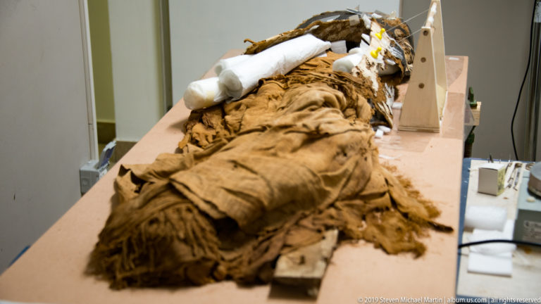Human mummy wrapped in thick brown linen lying on a conservation support with white padding near the head.