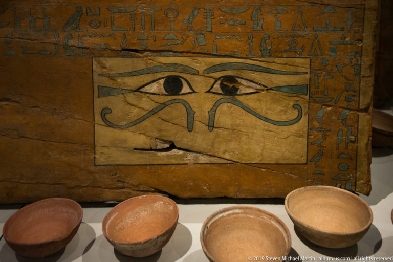 Painted wooden panel with large paired Eye of Horus motifs and hieroglyphs, displayed behind several pottery bowls.