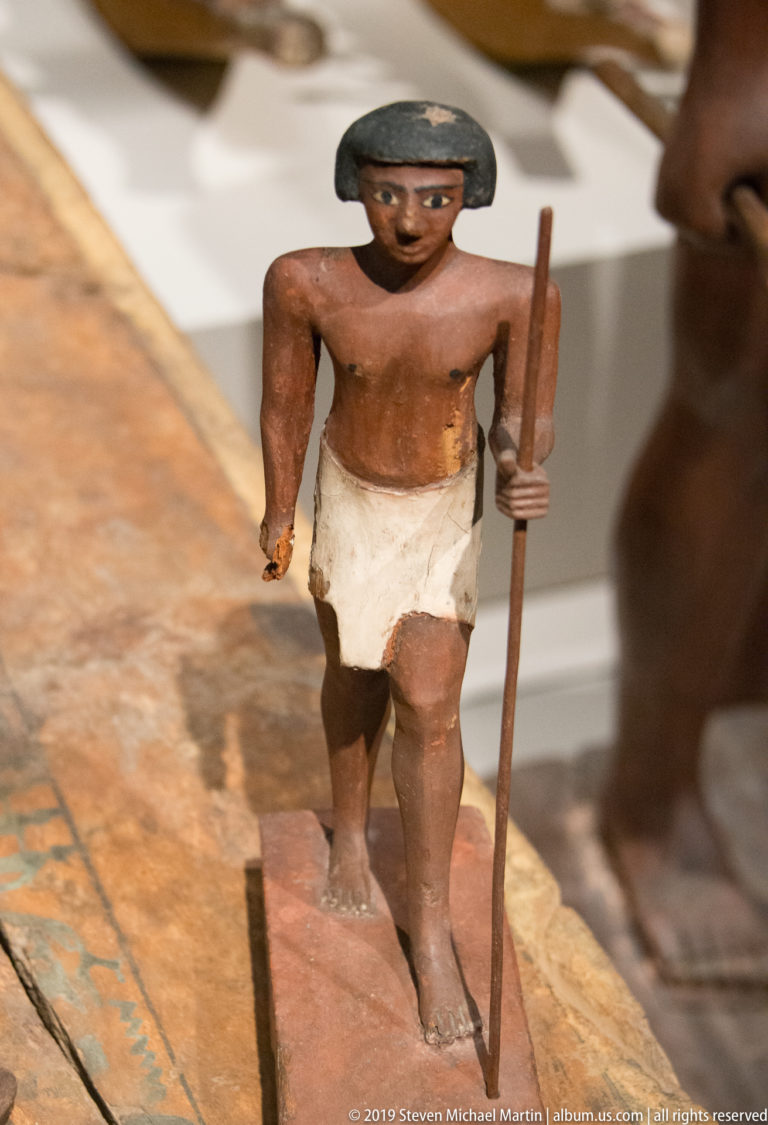 Small painted wooden Egyptian male figure with dark wig, white kilt, and long staff, standing with one foot forward.