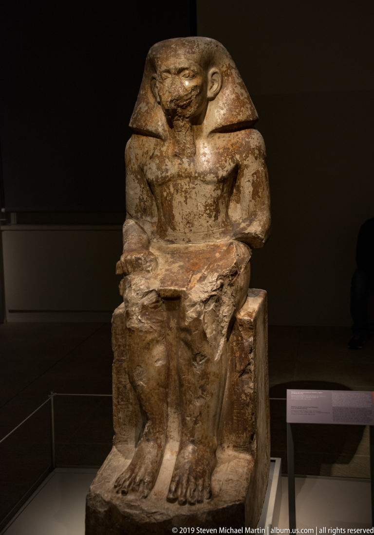 Stone statue of a seated Egyptian man wearing a broad wig or headdress, with hands resting across his lap.