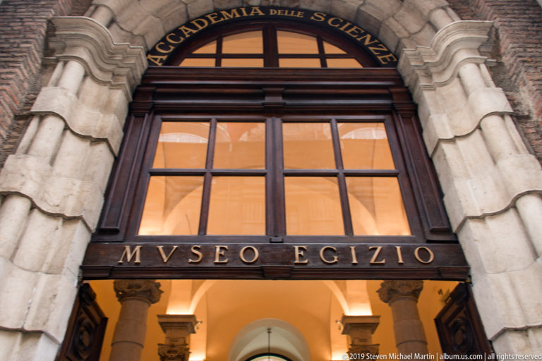 Historic stone portal with “MVSEO EGIZIO” inscription beneath tall windows and an arched institutional sign above.