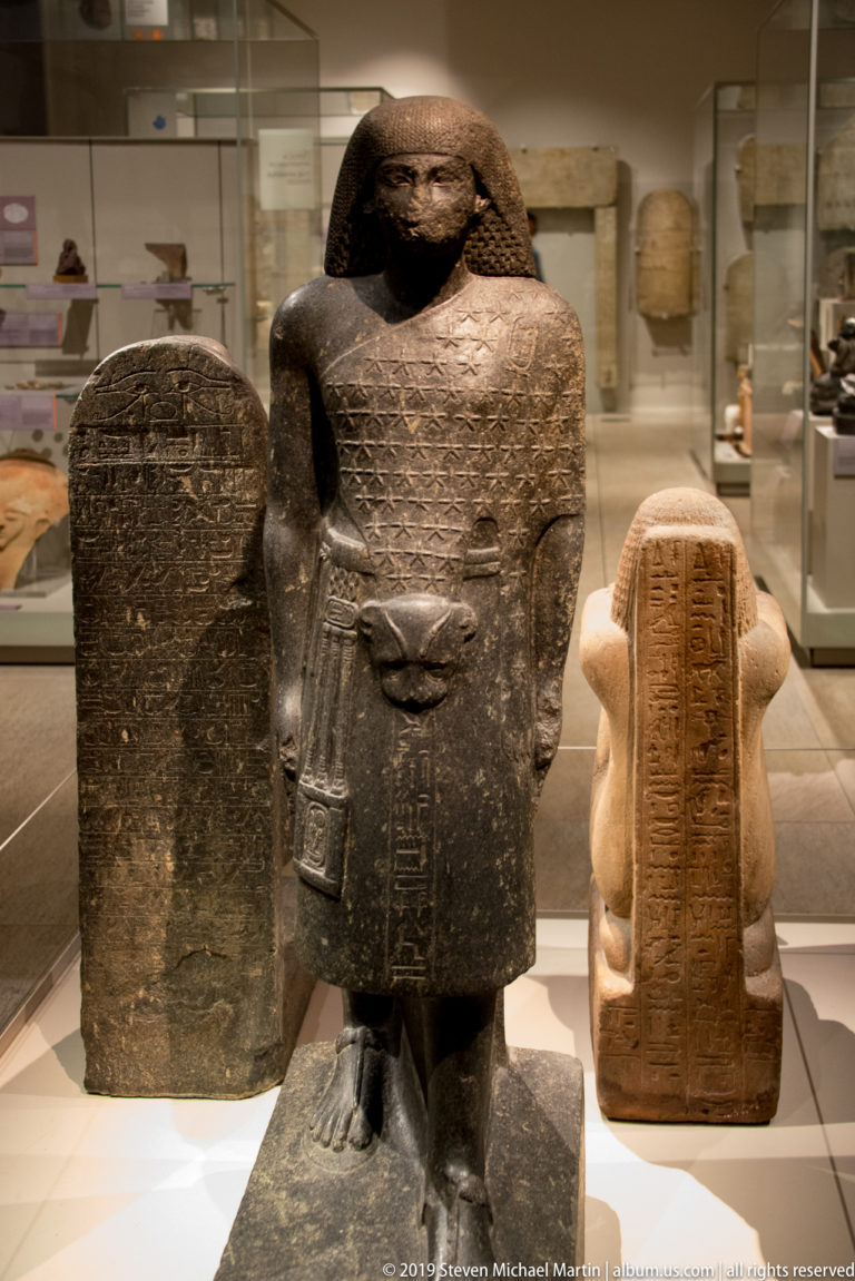 Dark Egyptian block statue of a man with head and feet emerging from a compact body covered in hieroglyphic inscriptions.