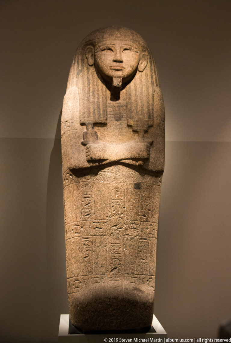 Nearly full-length granite anthropoid sarcophagus with broad wig, crossed arms, and dense hieroglyphic inscription across the lower body.
