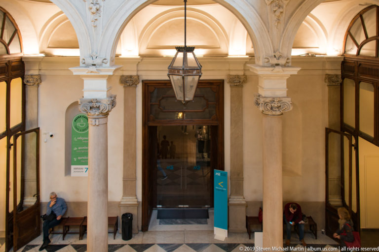 Arcaded museum hall with pale columns, carved capitals, rounded arches, hanging lantern, dark wood doors, and seated visitors.