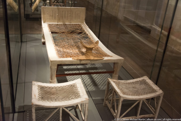 Wooden bed of Kha with woven plant-fiber surface, displayed with low woven stools from Tomb TT8 at Deir el-Medina.