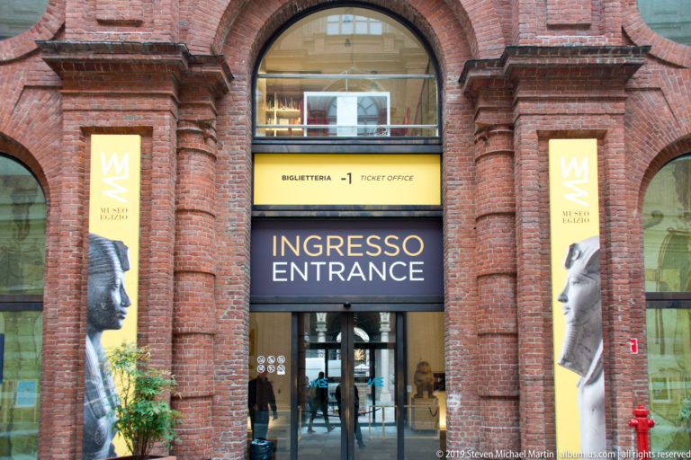 Main Museo Egizio entrance in a red-brick arched façade, with bilingual entrance sign, ticket office, and yellow museum banners.