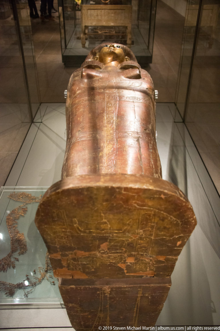 Head-end view of a reddish Egyptian anthropoid coffin showing the modeled face, chest, shoulders, and broad edge of the lower case.