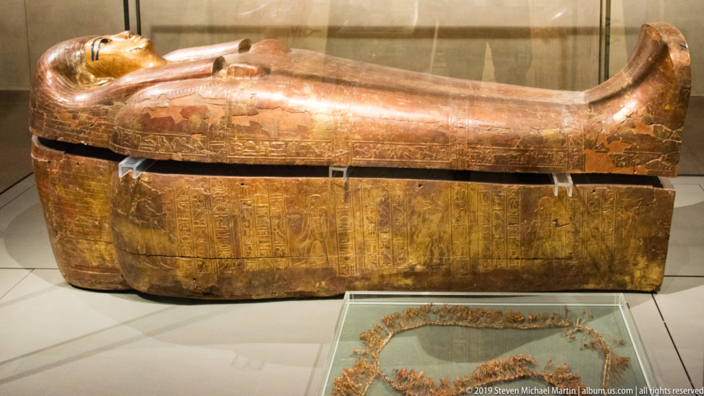 Reddish-brown anthropoid coffin above a rectangular hieroglyph-covered case, with a separate decorative object displayed in front.