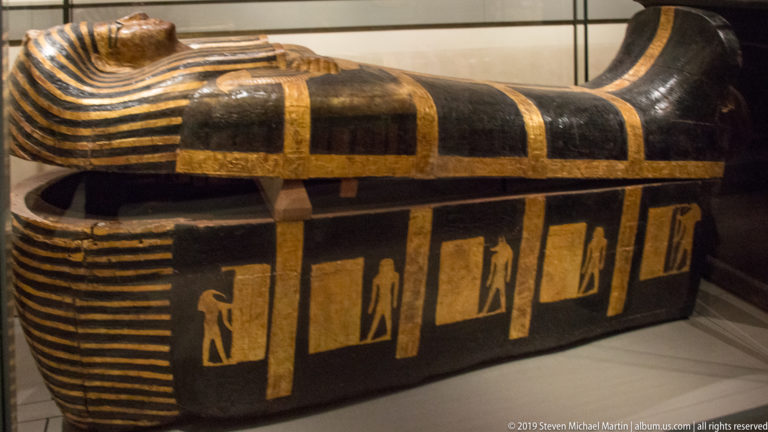 Side view of a black Egyptian anthropoid coffin with broad gold bands, striped ends, and small standing figures in rectangular panels.