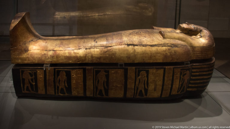 Horizontal black-and-gold anthropoid coffin with gilded upper body, striped ends, and standing figures painted in side panels.