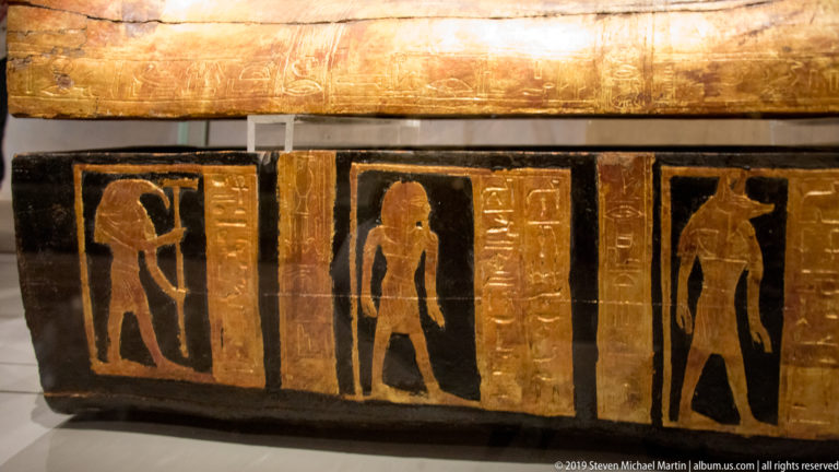Black-and-gold Egyptian coffin side with standing figures in vertical panels separated by narrow hieroglyphic columns.