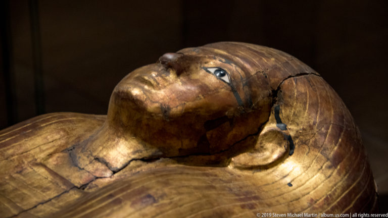 Cracked gilded Egyptian coffin face and upper body with painted eye, broad wig, and visible surface losses.