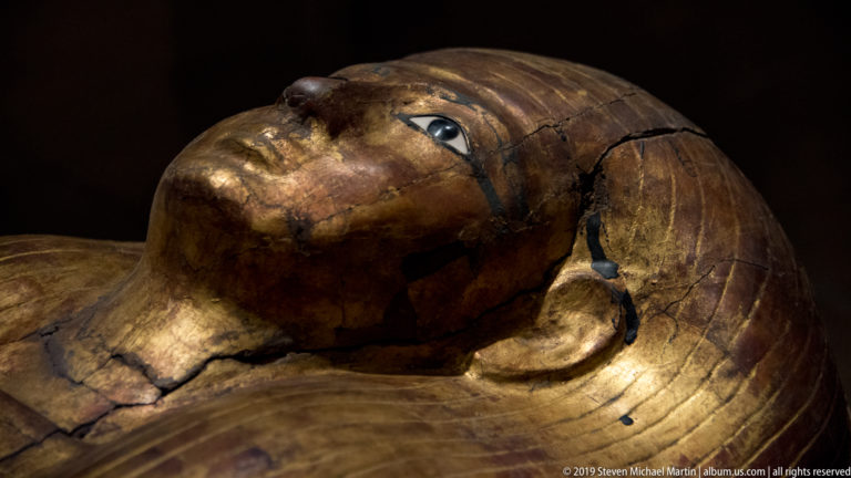 Angled close view of a worn gilded Egyptian coffin face with outlined eye, extensive cracking, and surface loss.