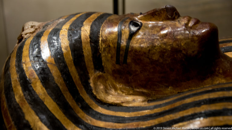 Profile close-up of an Egyptian anthropoid coffin face with painted eye, dark cosmetic line, and alternating black-and-gold wig bands.
