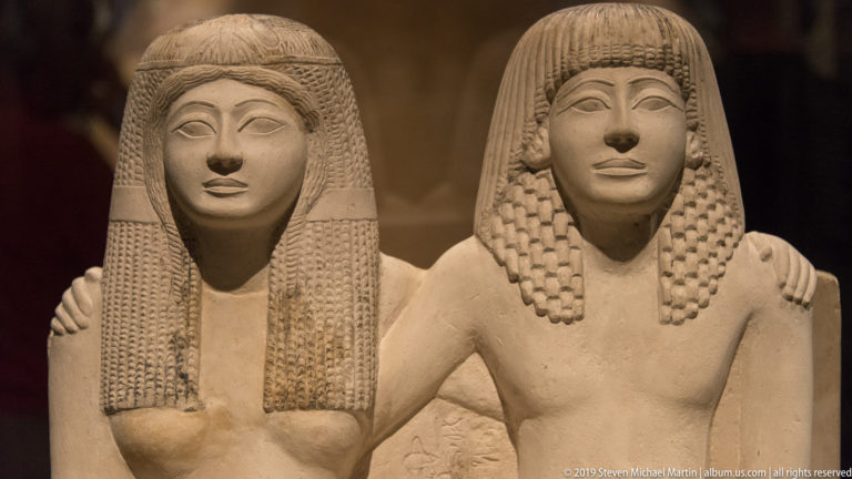 Paired Egyptian stone portrait of a woman and man with elaborate wigs, idealized faces, and an arm linking the two figures.