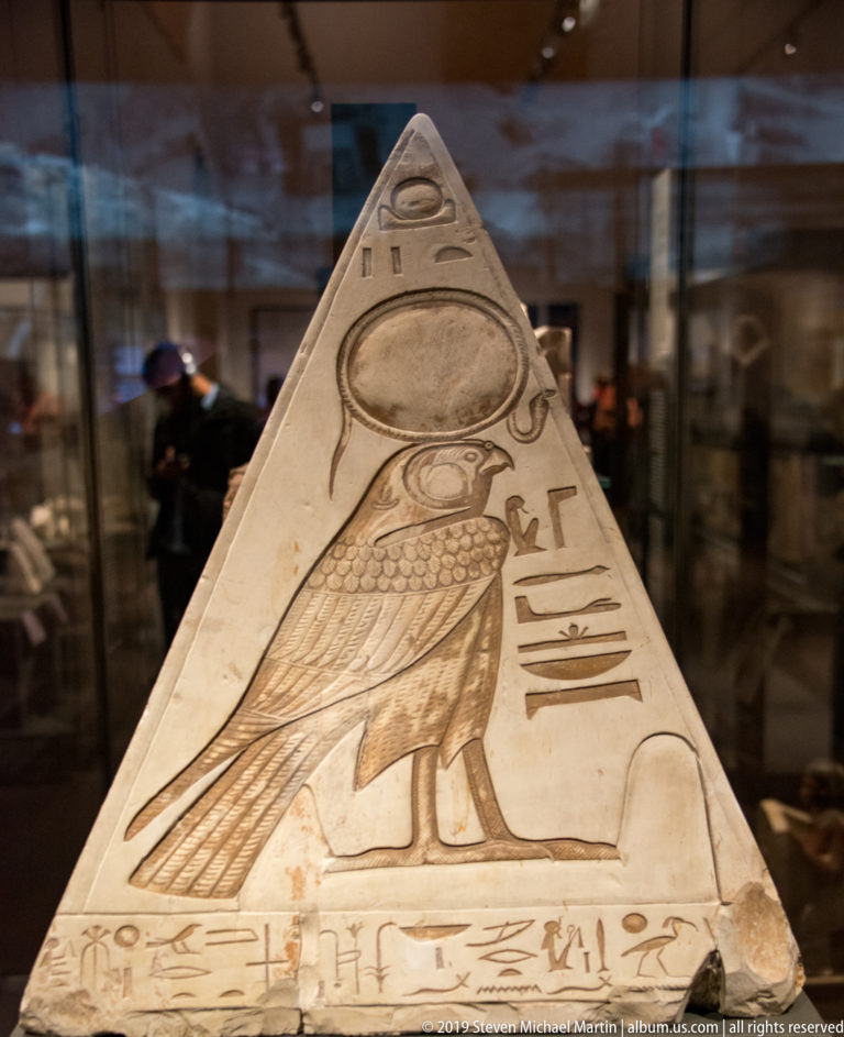Triangular Egyptian pyramidion carved with a falcon beneath a solar disk and hieroglyphic inscriptions along the face and base.