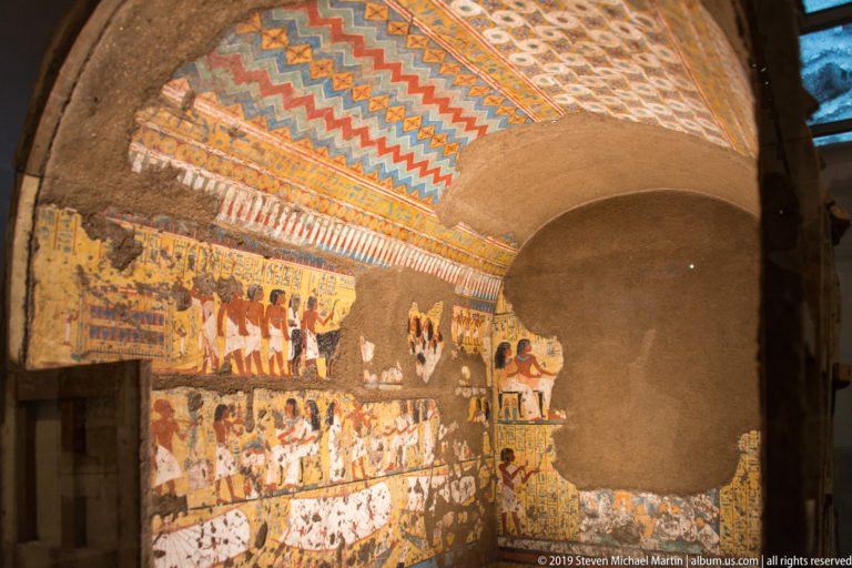 Egyptian painted tomb chamber with a large arched recess, colorful geometric ceiling, and processional wall scenes.