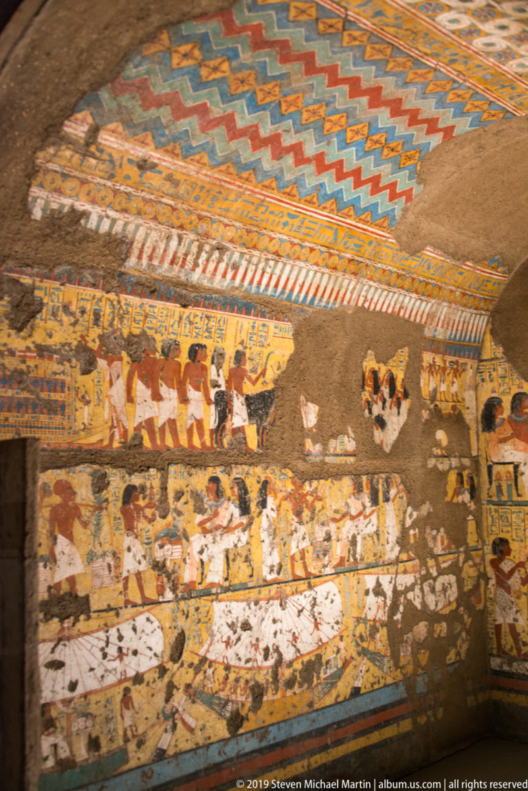 Painted Egyptian tomb wall with processional figures beneath a brightly colored geometric ceiling, with areas of plaster loss.