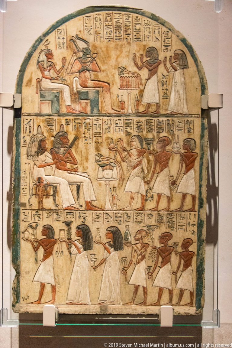 Rounded-top Egyptian funerary stela with hieroglyphic text and several registers of seated, standing, and kneeling figures.