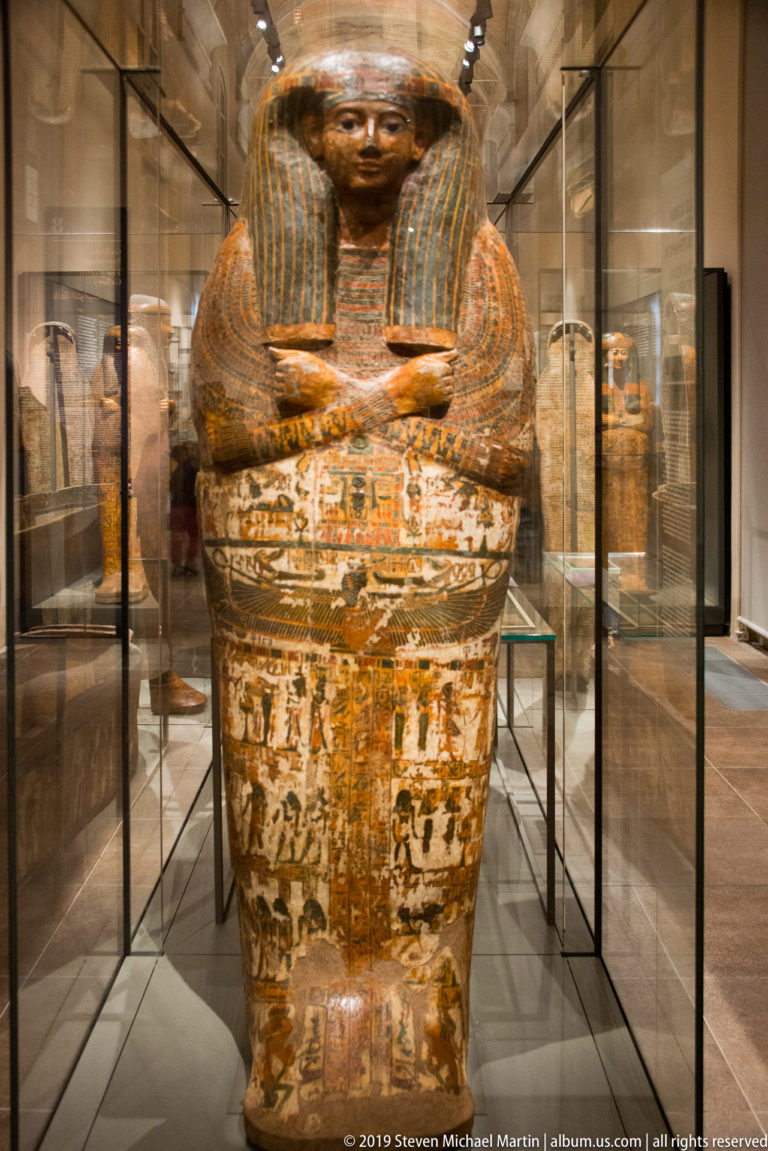 Upright painted Egyptian anthropoid coffin with dark wig, modeled face, hieroglyphic bands, and dense multicolored decoration.